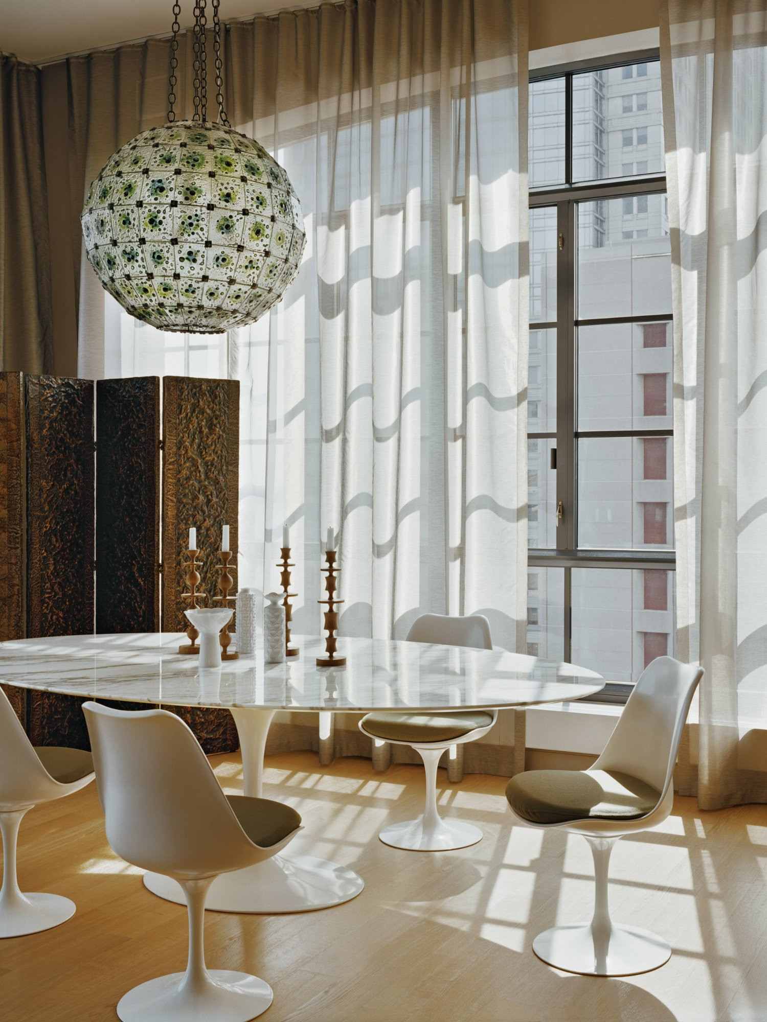Chaises Tulipe blanches sans accoudoirs et coussins verts autour d’une table ovale devant de grandes fenêtres.